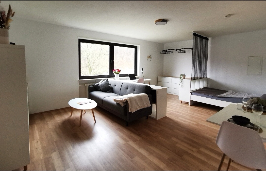 A spacious living area is presented with a gray sofa and a light wood coffee table. Large windows provide natural light, and a bed is positioned in the background. A minimalist dresser and a coat rack are visible, contributing to the apartment's streamlined design.