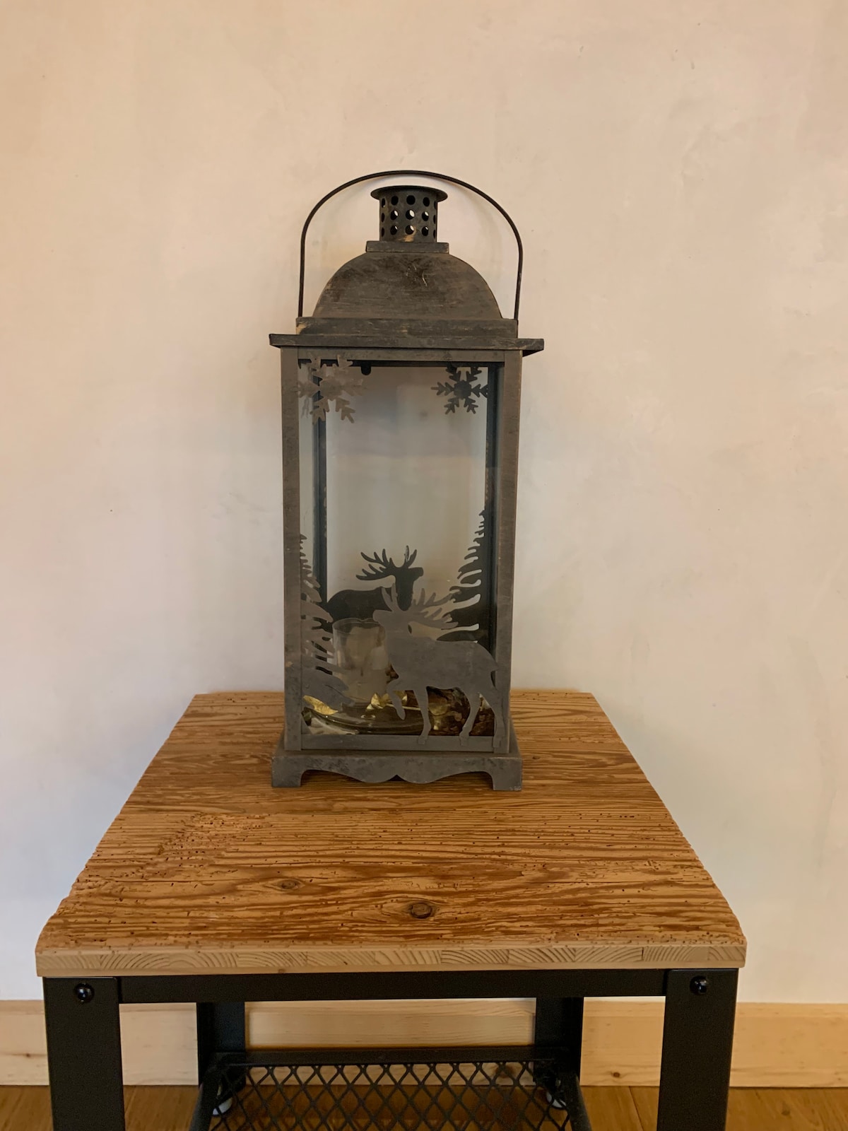 An ornamental lantern is displayed on a wooden table, featuring intricate cut-outs of forest scenes with deer. The lantern's metal frame is complemented by a rustic finish, adding a charming decorative touch to the space.