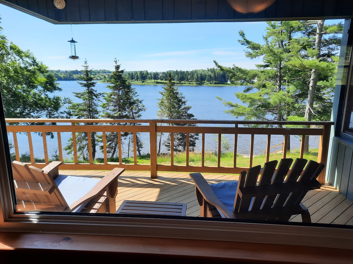 A wooden deck is visible through a large window, featuring two Adirondack chairs positioned towards the river. The scenery includes trees lining the shore, with calm water reflecting the blue sky.