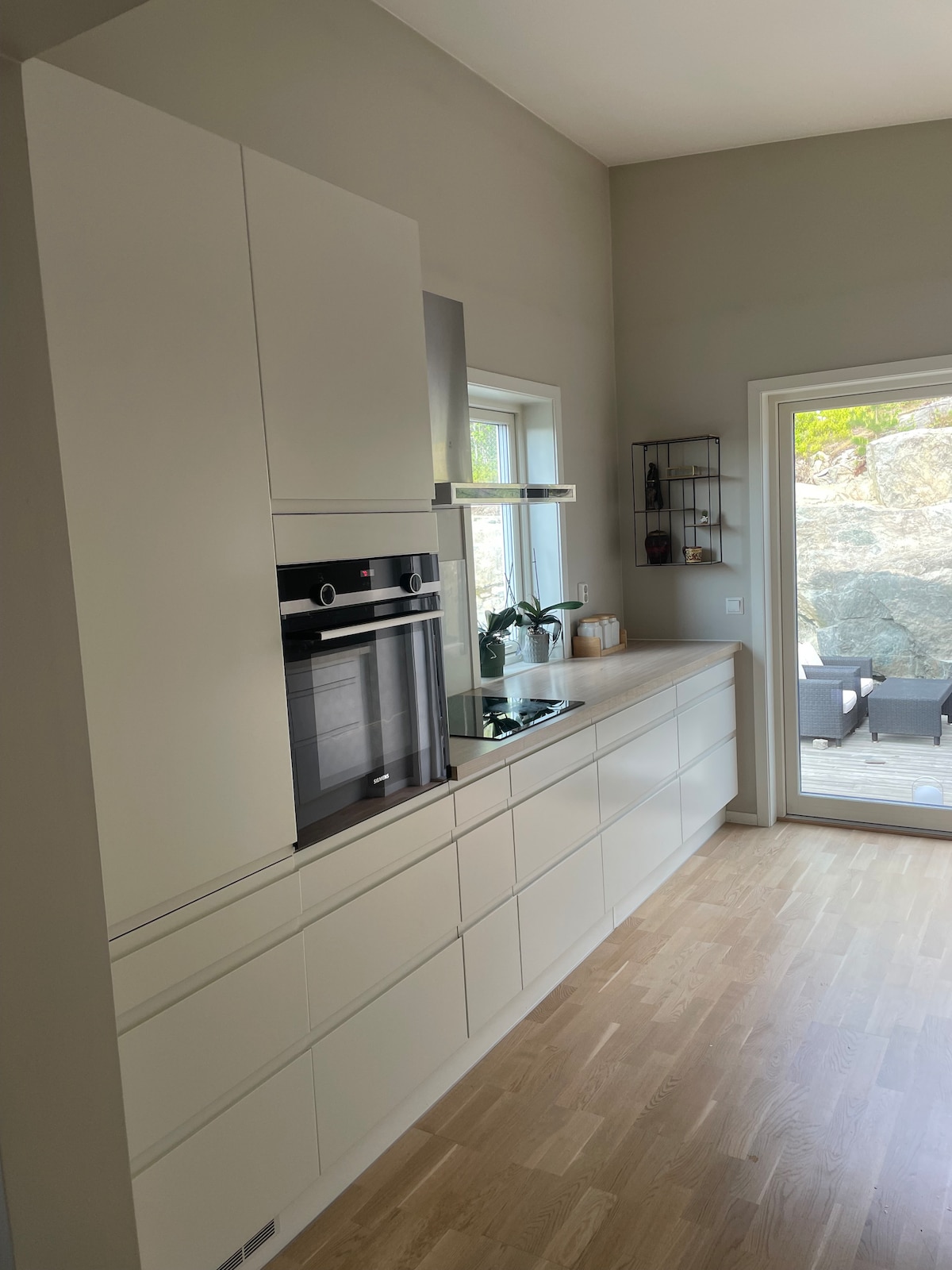 A modern kitchen features sleek white cabinetry and integrated appliances, including an oven and cooktop. Natural light brightens the space through a large window, with a glimpse of a patio area visible outside, complemented by greenery.