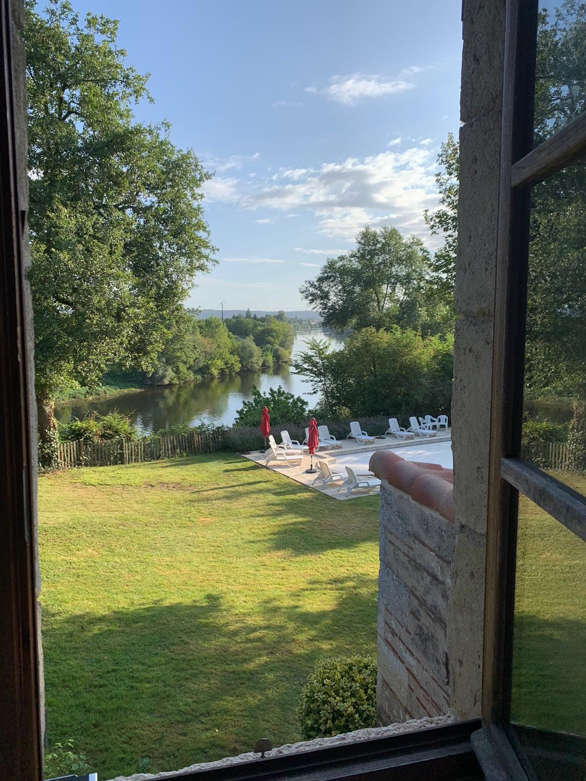 A view from an interior window reveals a lush green lawn leading to the Dordogne river. The pool area is visible, featuring white lounge chairs and parasols, with trees framing the scene under a clear blue sky.