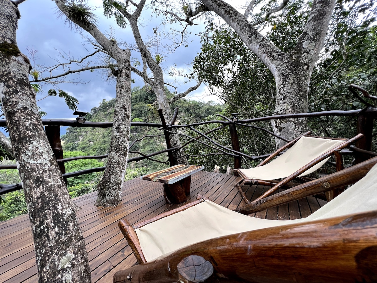 A wooden deck is nestled among trees, featuring two lounge chairs positioned for relaxation. A small table sits between them, surrounded by lush greenery and the natural landscape beyond. The space invites guests to enjoy tranquility and the scenic views.