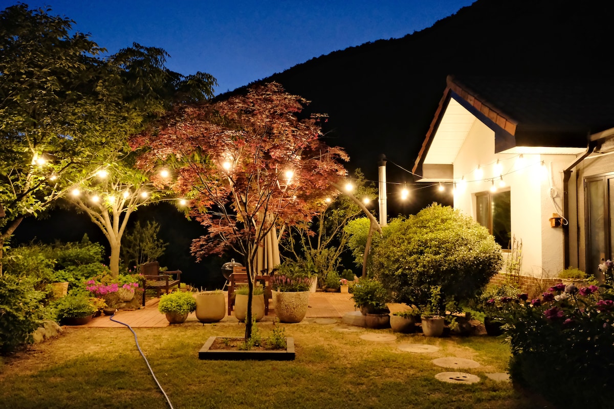 An outdoor space is softly lit by string lights, illuminating various plants and garden features. A small tree adds a splash of color, while a patio area includes seating and markers of lush greenery. The backdrop reveals a mountainous silhouette under the evening sky.