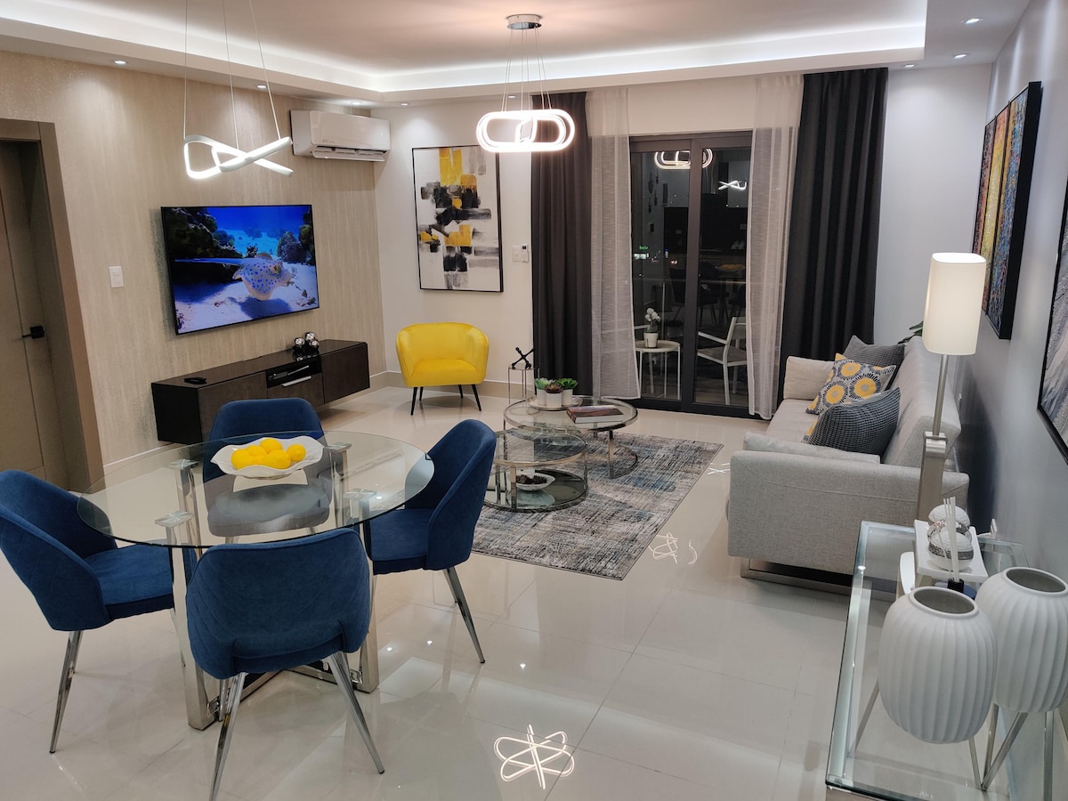 A modern living area features a plush gray sofa paired with accent chairs in blue. A glass dining table is set for four, with a decorative centerpiece. Soft lighting is provided by contemporary pendant fixtures, and large windows invite natural light, offering a view of the outside.