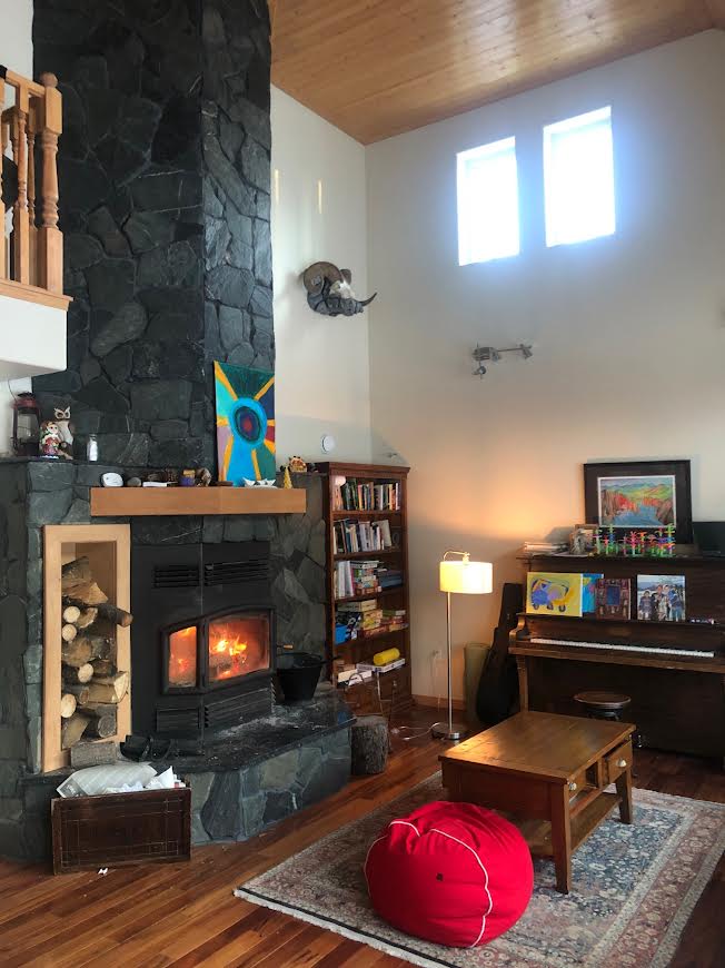 A cozy family room features a stone fireplace with a flickering flame, surrounded by wooden panels and a bookshelf filled with books. A lamp provides soft light near a piano, while a small table and a decorative red pouf complete the welcoming space.