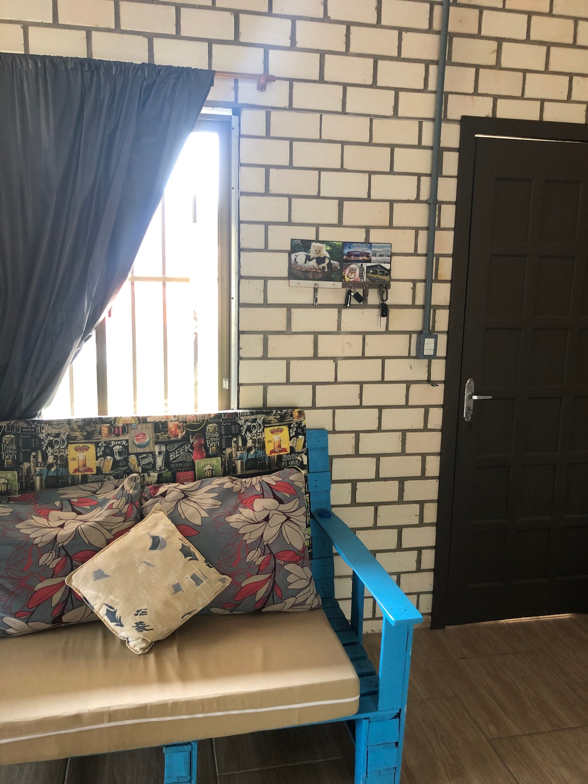 A corner seating area is presented with a wooden bench adorned with floral-patterned cushions. A light-colored wall features exposed brick elements, while a dark curtain drapes over the window, allowing filtered light to enter. Nearby, photographs are displayed on the wall.