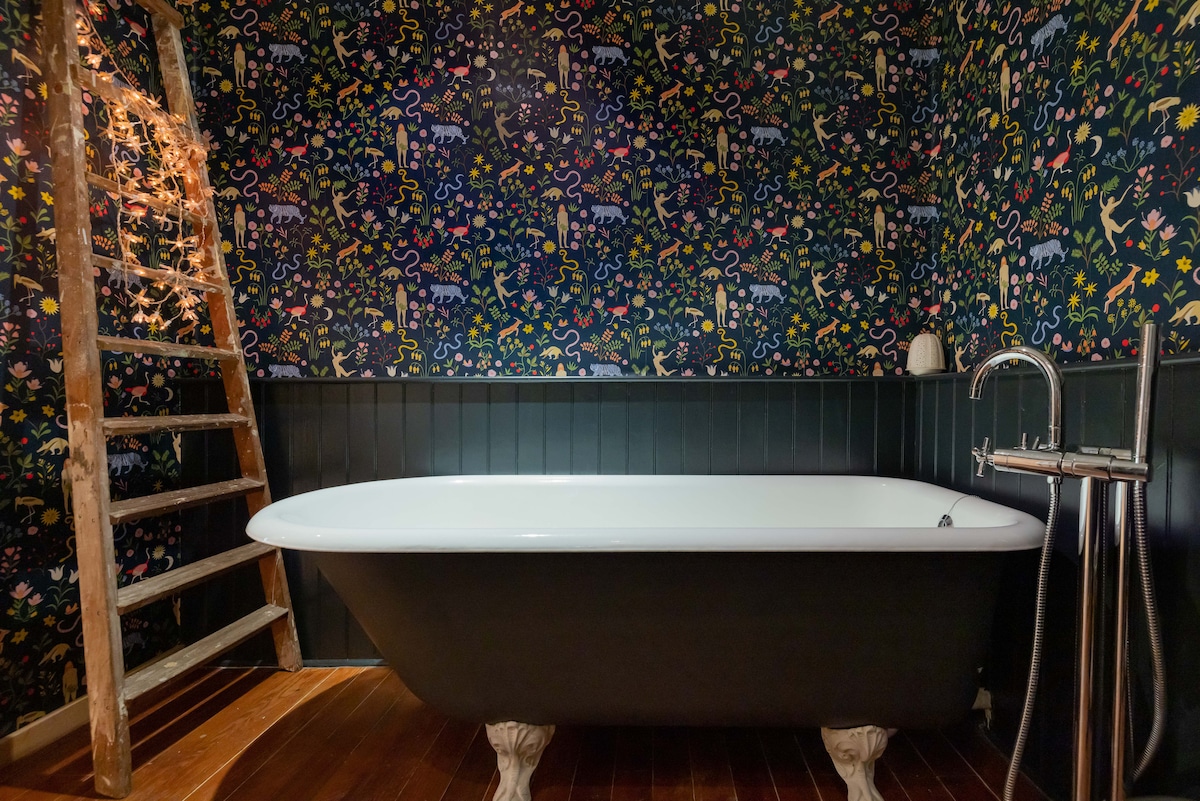 An elegant roll-top bathtub with a black exterior is centered against a vibrant, patterned wallpaper featuring whimsical designs. A wooden ladder rests against the wall, adorned with string lights. Dark wood flooring complements the overall aesthetic and adds warmth to the space.