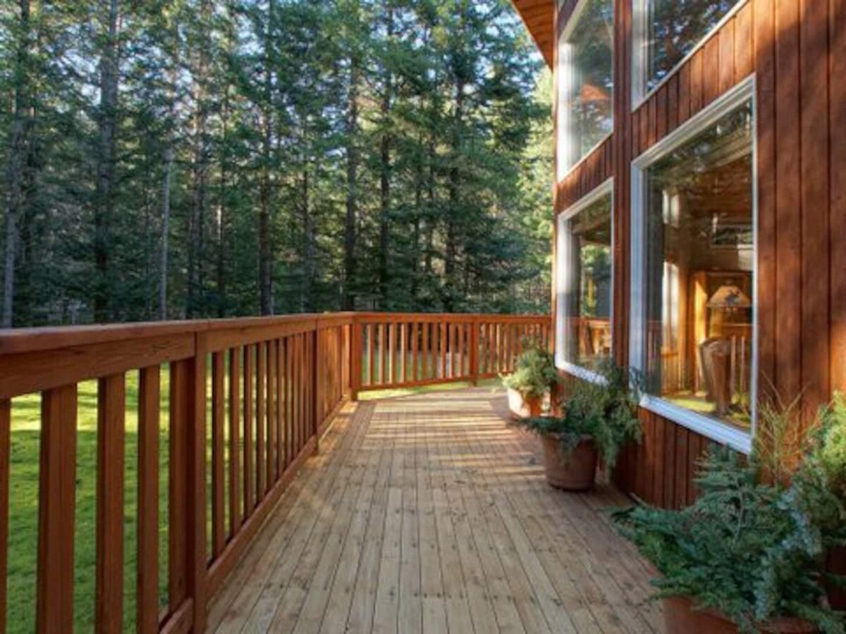 A wooden deck extends alongside a cabin, surrounded by tall trees. Potted plants are placed at intervals along the railing, and sunlight casts gentle shadows on the deck's surface, enhancing the natural ambiance.
