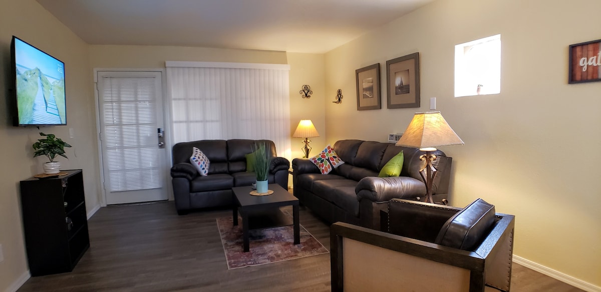 The living room presents a welcoming space with two comfortable leather sofas and a matching armchair. A coffee table is centered on a decorative rug. A TV mounted on the wall provides entertainment options. Soft lighting is provided by lamps, enhancing the room's coziness.