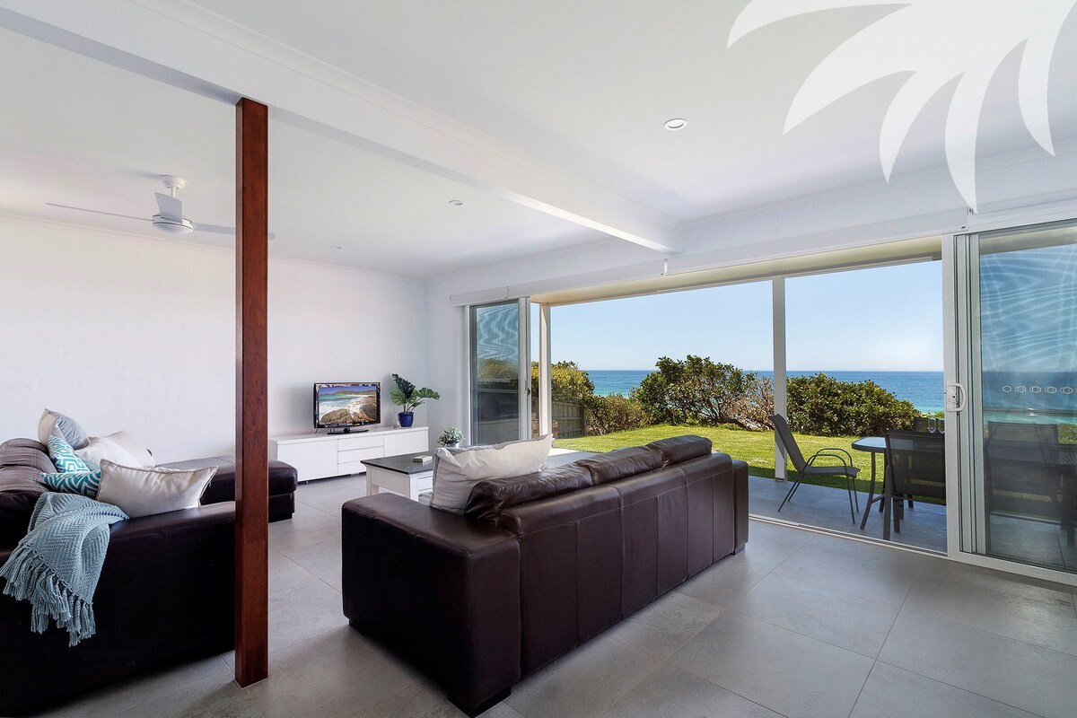 A modern living area features a dark leather sectional sofa, complemented by a textured throw. Large glass doors provide ocean views and open to an outdoor seating area. A ceiling fan and a wall-mounted television enhance the space's functionality.