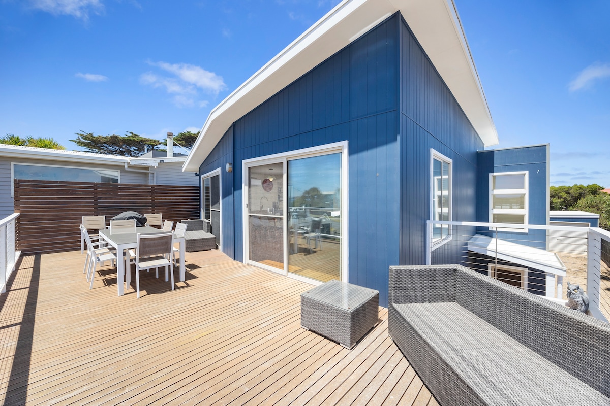 A spacious outdoor deck features a comfortable seating area with a modern grey couch and a dining table with several chairs. Large sliding doors provide access to the interior, while the surrounding fence ensures privacy. Bright blue skies enhance the inviting atmosphere.