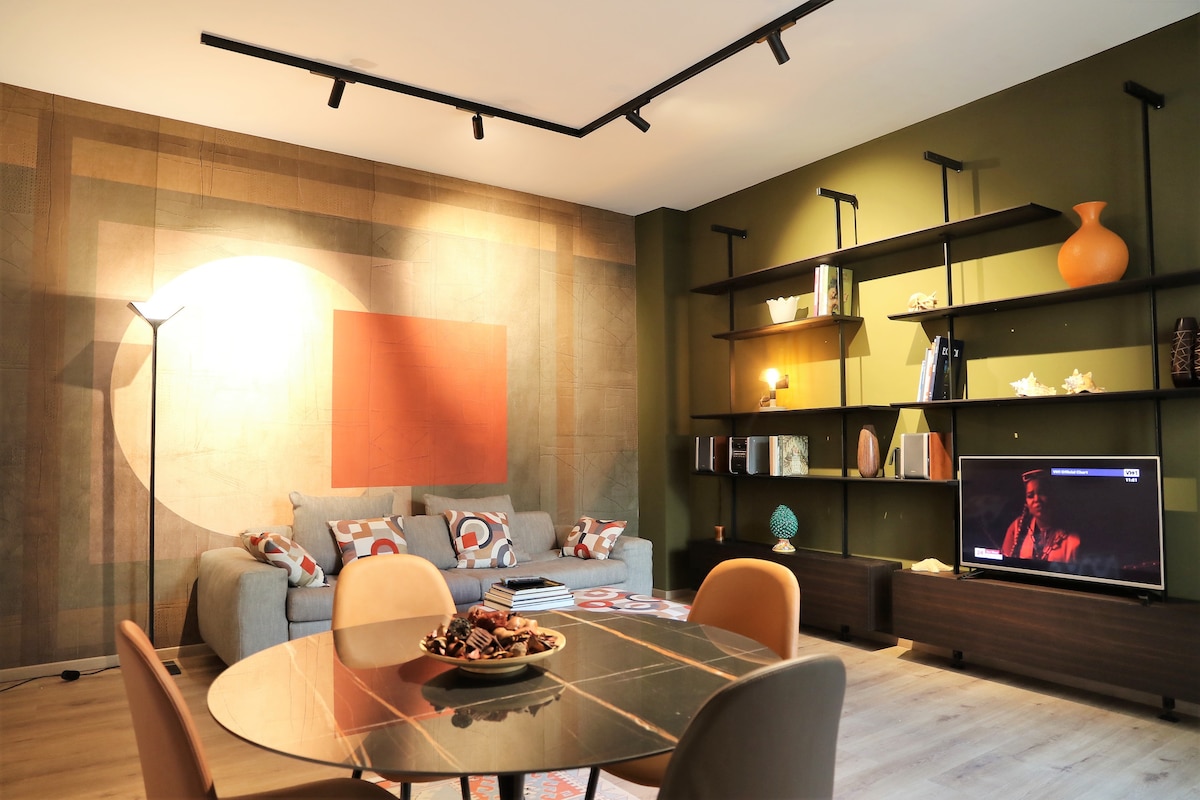 A modern living area features a comfortable seating arrangement with decorative cushions. A round dining table sits in the foreground, complemented by stylish chairs. A flat-screen TV is positioned on a dark wooden console, while shelves display decorative items against a textured wall.