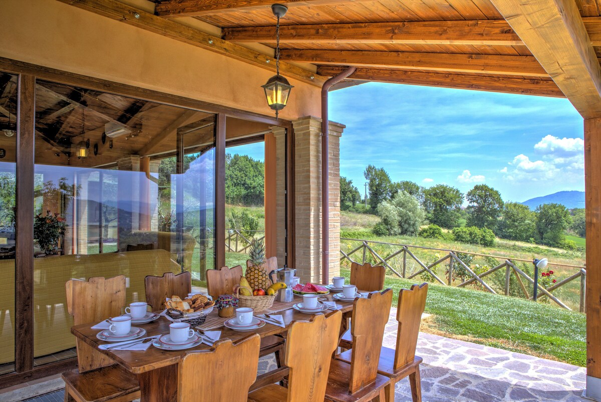 An outdoor dining area is showcased under a wooden pergola, featuring a large table set for breakfast. Multiple wooden chairs surround the table, which is adorned with plates, cups, and an assortment of fruits. Scenic hills and lush greenery are visible in the background.