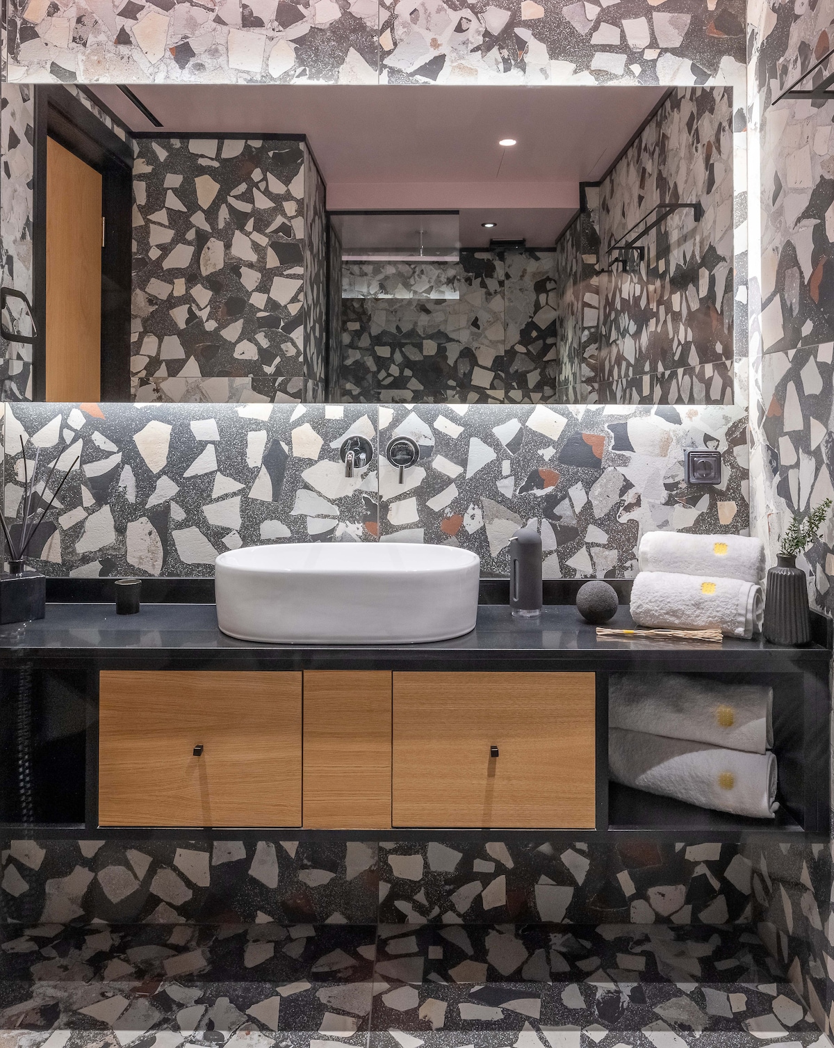 The modern bathroom features a sleek countertop with a minimalist sink, surrounded by textured stone walls. Built-in wooden cabinets provide storage, and neatly rolled towels are placed nearby. Subtle lighting enhances the contemporary design.