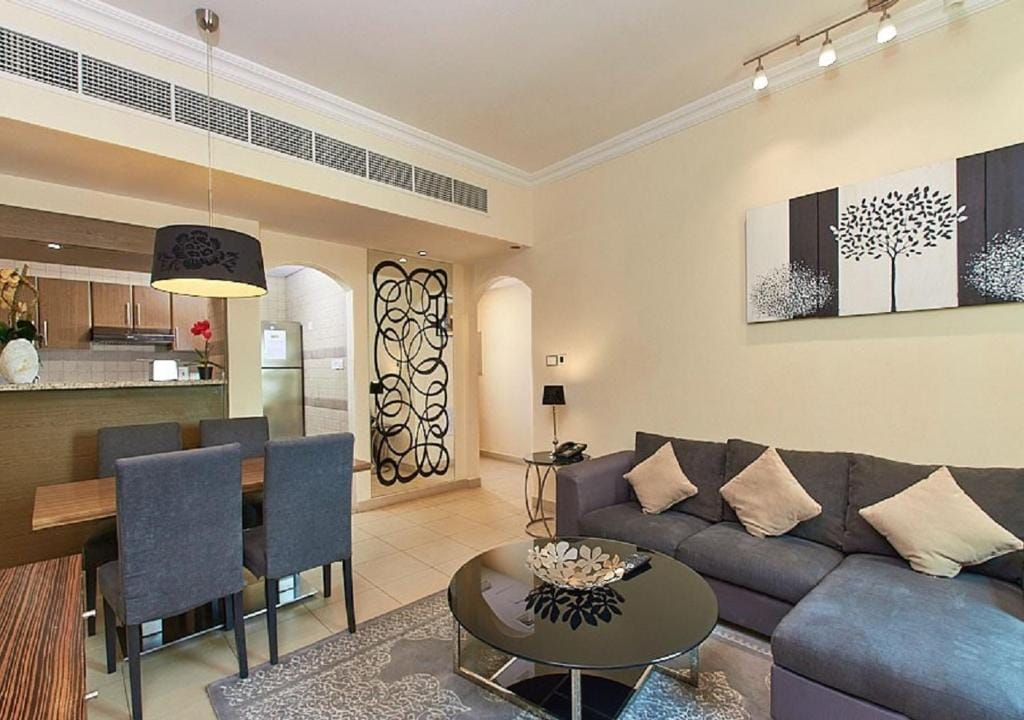 A cozy living area is shown, featuring a modern sectional sofa with decorative pillows and a sleek circular coffee table. A dining table with four chairs is visible, complemented by ambient lighting and contemporary artwork on the wall.