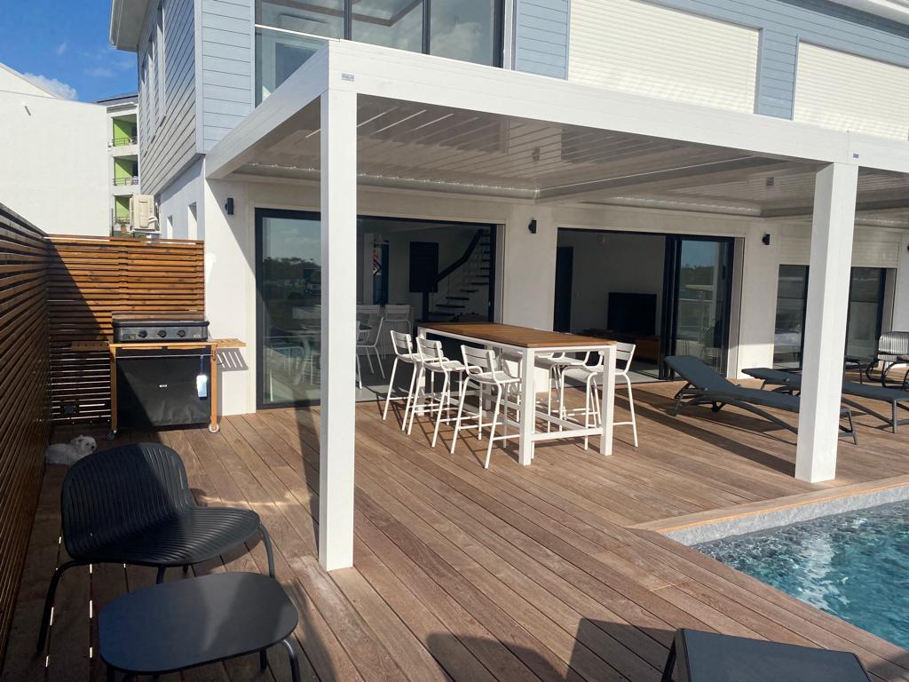 An outdoor patio area features a long wooden dining table surrounded by white chairs. A barbecue grill is positioned nearby on the wooden deck, which is bordered by sun loungers and framed by sleek glass doors leading to the interior space.
