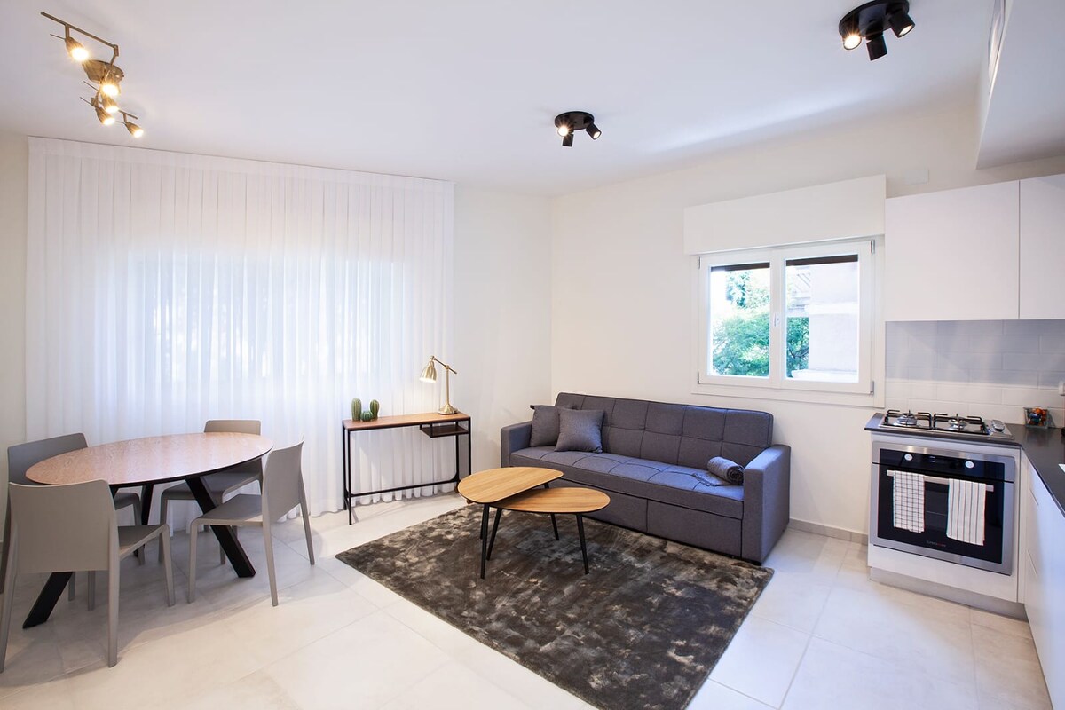 A modern living space features a comfortable gray sofa and two coffee tables on a soft area rug. A dining table with four chairs is positioned by the window, while natural light brightens the space. A small lamp and a side table add subtle decor.