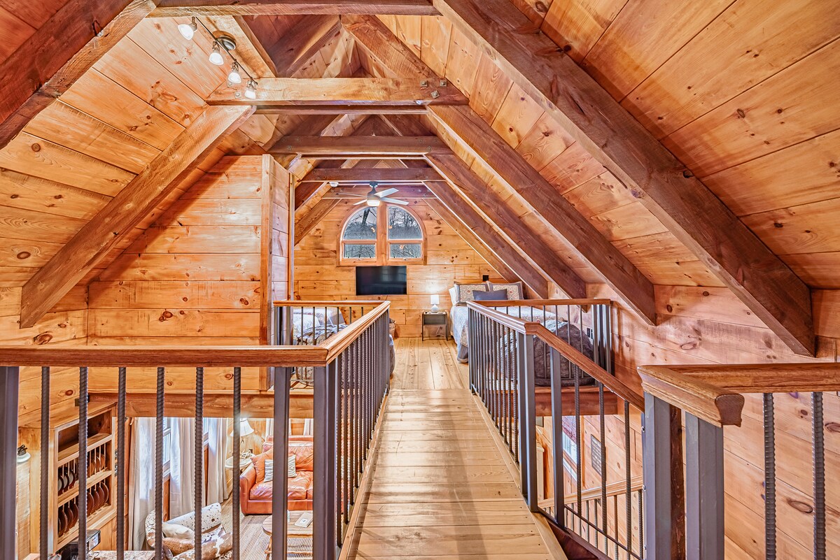 The spacious loft area is characterized by wooden beams and an open layout. The balcony railings overlook the living area below. Natural light floods in through an arched window, and a Smart TV is visible at the far end of the loft.