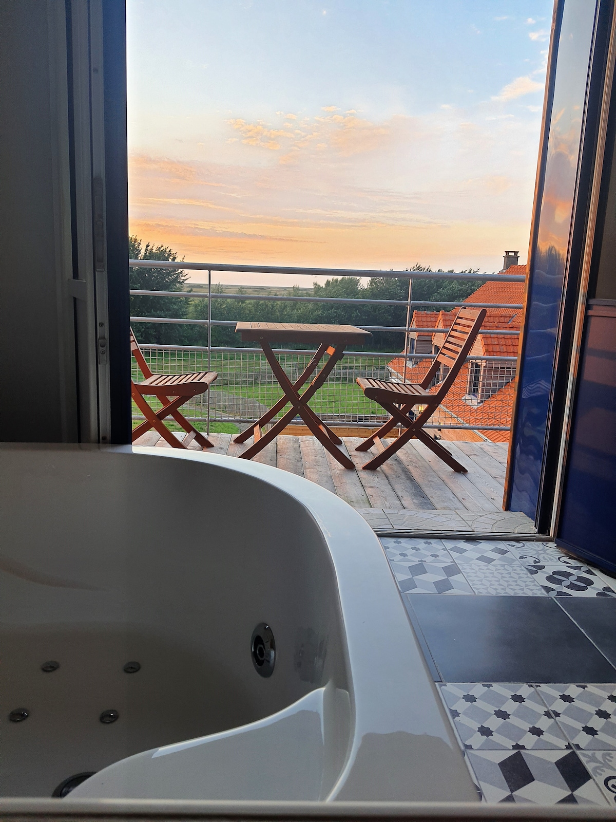 A cozy bathroom corner is featured, showcasing a modern bathtub positioned beside an open door. Outside, a balcony is visible with two wooden chairs and a small table, set against a backdrop of a colorful sunset and green surroundings.