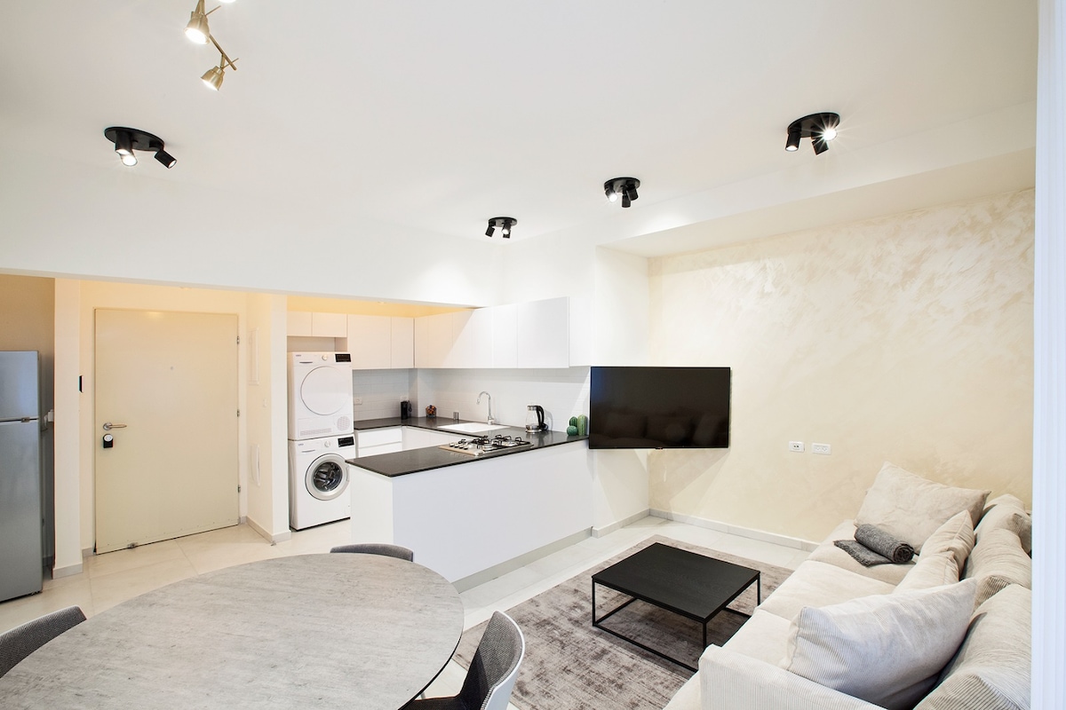 A modern kitchen area is visible, featuring sleek white cabinetry and black countertops. A dining table accommodates several seats, and a comfortable sofa is positioned nearby. Bright overhead lighting enhances the clean and fresh design, while a large wall-mounted television adds functionality.