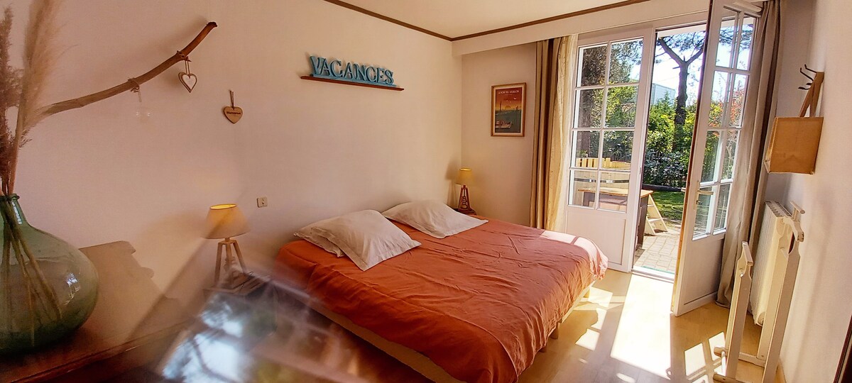 A spacious sleeping area features a double bed with light orange bedding. Soft lighting is provided by two bedside lamps. A set of glass doors opens to a private terrace surrounded by greenery, inviting natural light into the room.
