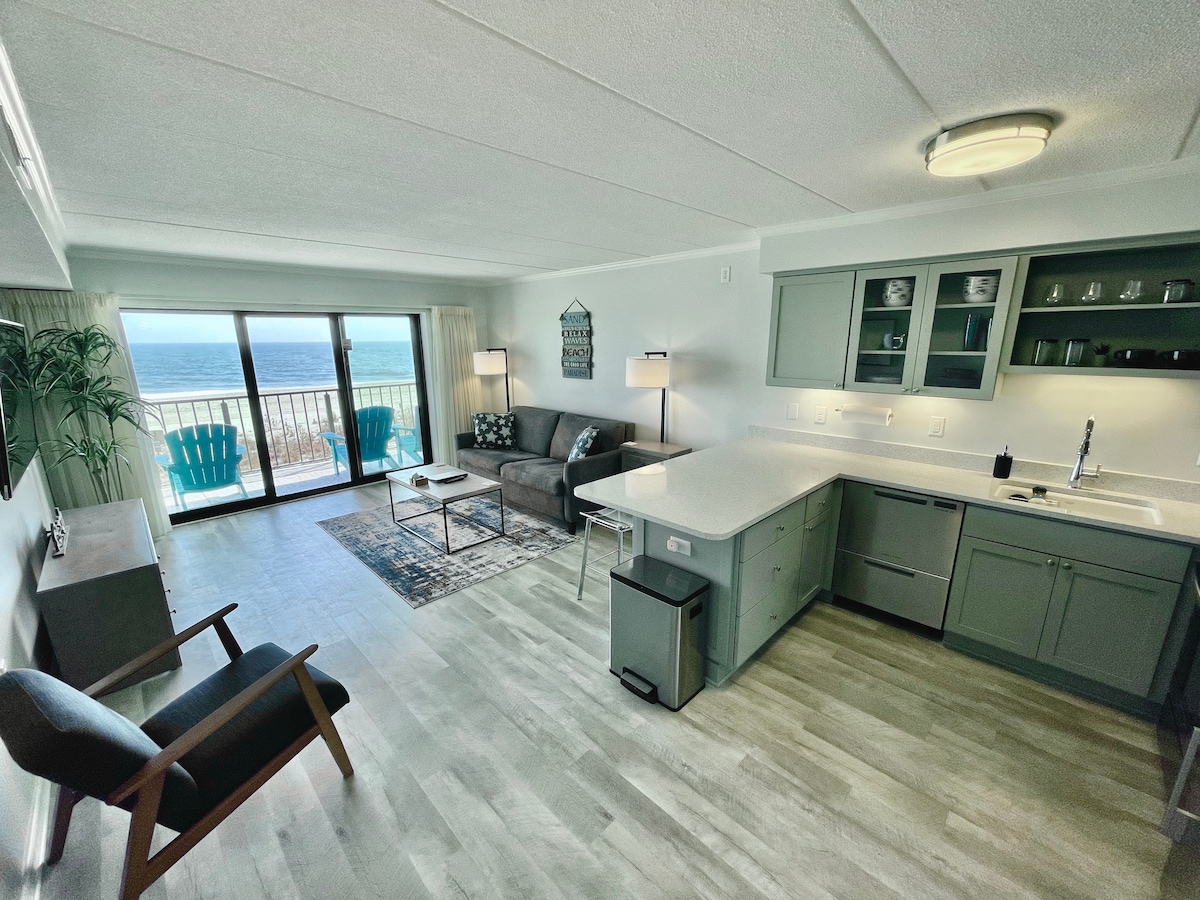 A spacious living area showcases a modern kitchen with sleek cabinetry and a cozy seating arrangement. Large glass doors provide a view of the ocean and lead to a private balcony. Decorative elements include a patterned rug and two vibrant blue chairs.