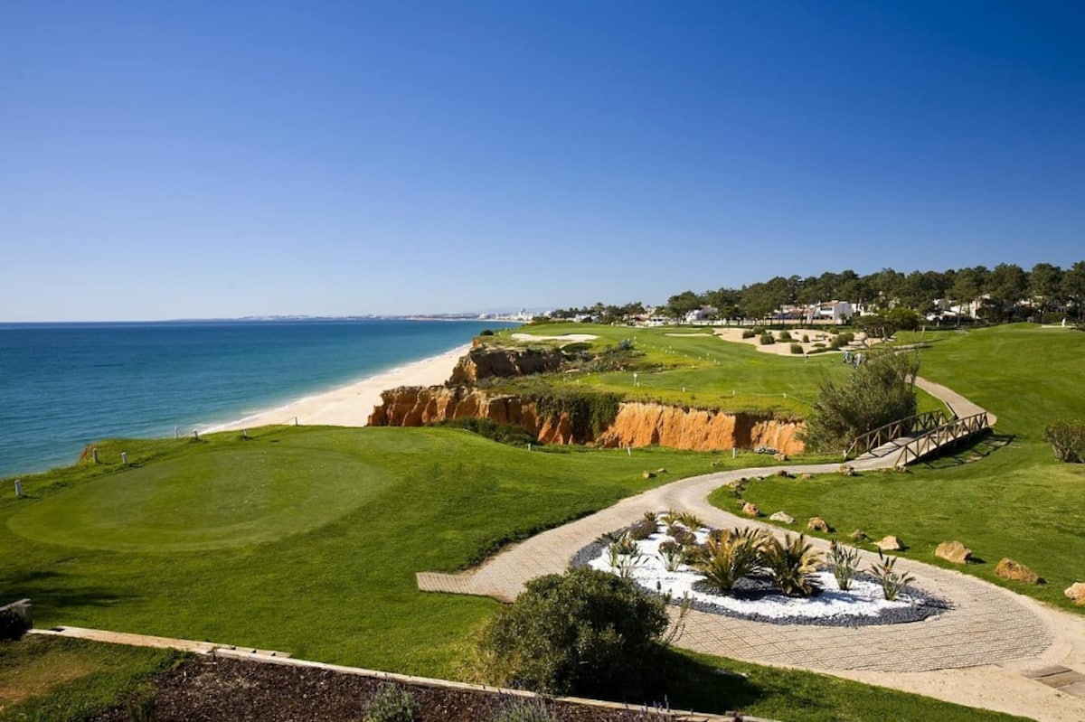 A scenic view of the coastline blends sandy beaches with lush green golf courses. The shoreline stretches towards the horizon under a clear blue sky. A landscaped pathway featuring rocks and plants leads through the foreground, enhancing the natural beauty of the surroundings.
