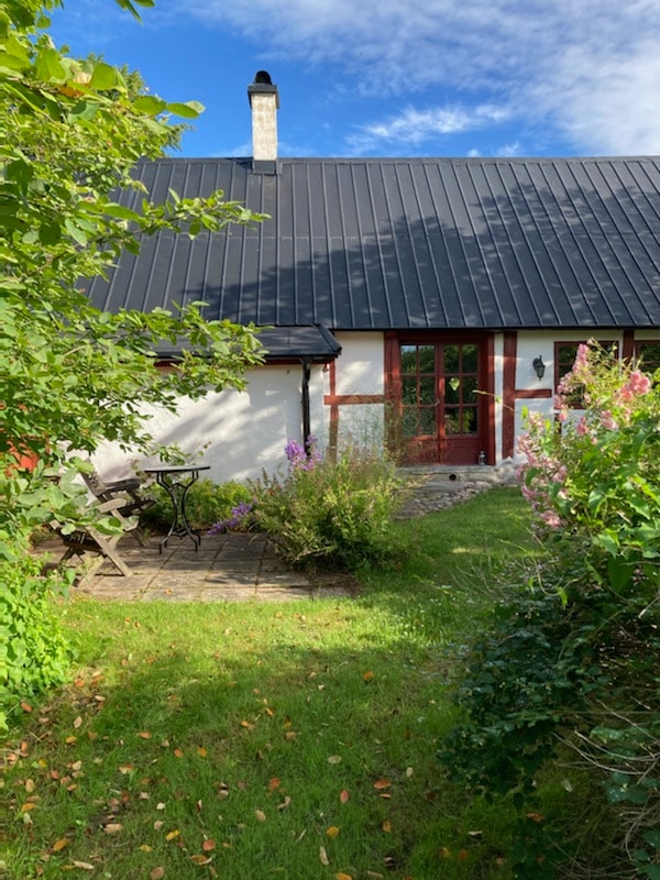A serene outdoor area is depicted, featuring a small stone patio with a bistro table and two chairs surrounded by lush greenery. Colorful flowers and an inviting garden create a peaceful atmosphere, while the building's traditional architecture is highlighted by a dark metal roof.
