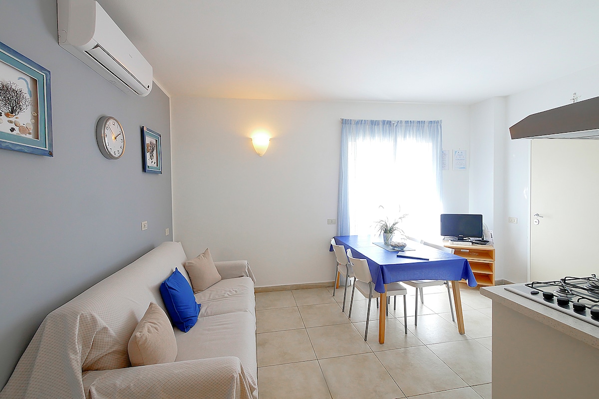 A bright living area features a white sofa adorned with blue cushions, complemented by a dining table set for four. Natural light fills the space through a large window with light curtains, while a wall clock and decorative frames add to the welcoming ambiance.