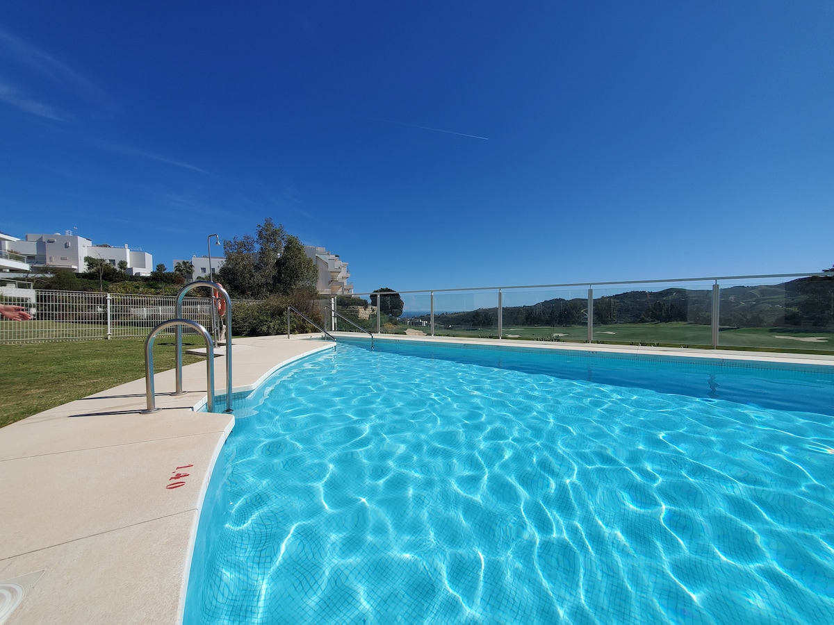The swimming pool is situated against a clear blue sky, showcasing sparkling water that reflects sunlight. A smooth deck surrounds the pool, with safety steps leading into the water. Lush greenery and distant mountains are visible in the background, enhancing the tranquil setting.