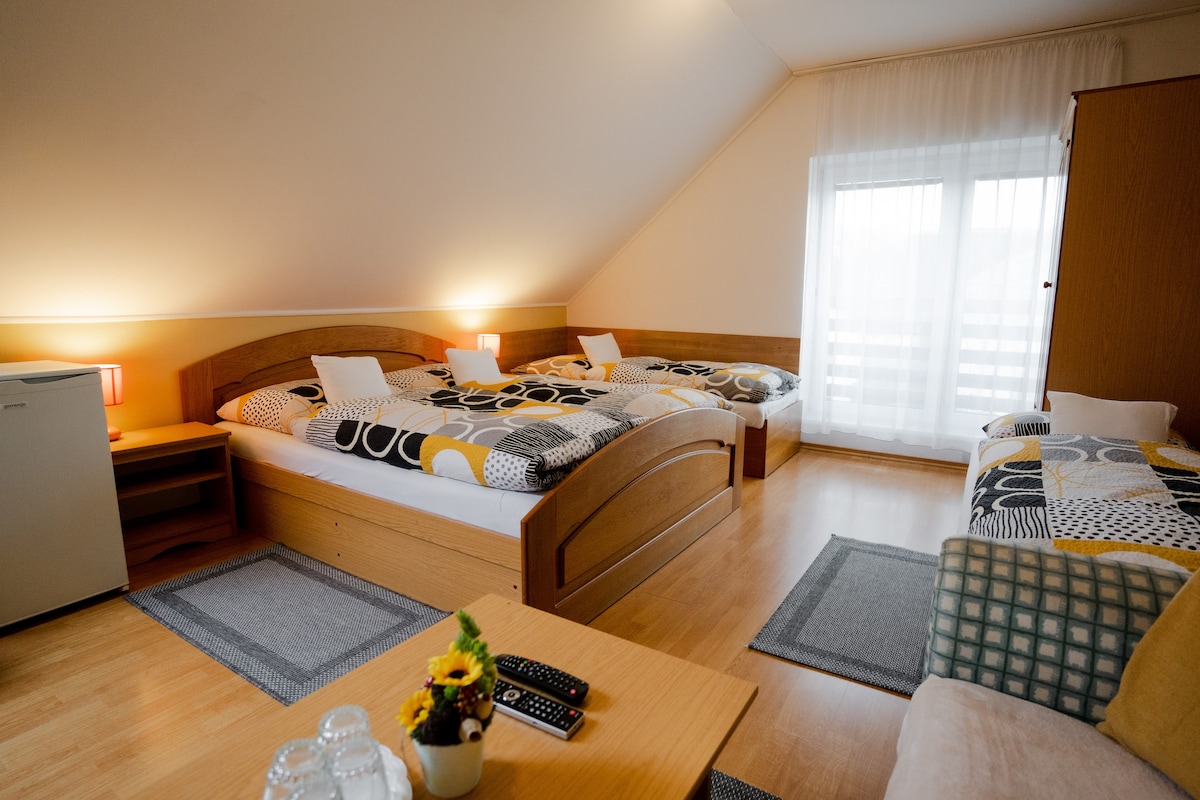 A spacious room features two wooden beds with patterned bedding and an additional single bed. A small table holds a remote control and a vase with sunflowers. A refrigerator and a wardrobe are present, with light filtering through sheer curtains covering the window.