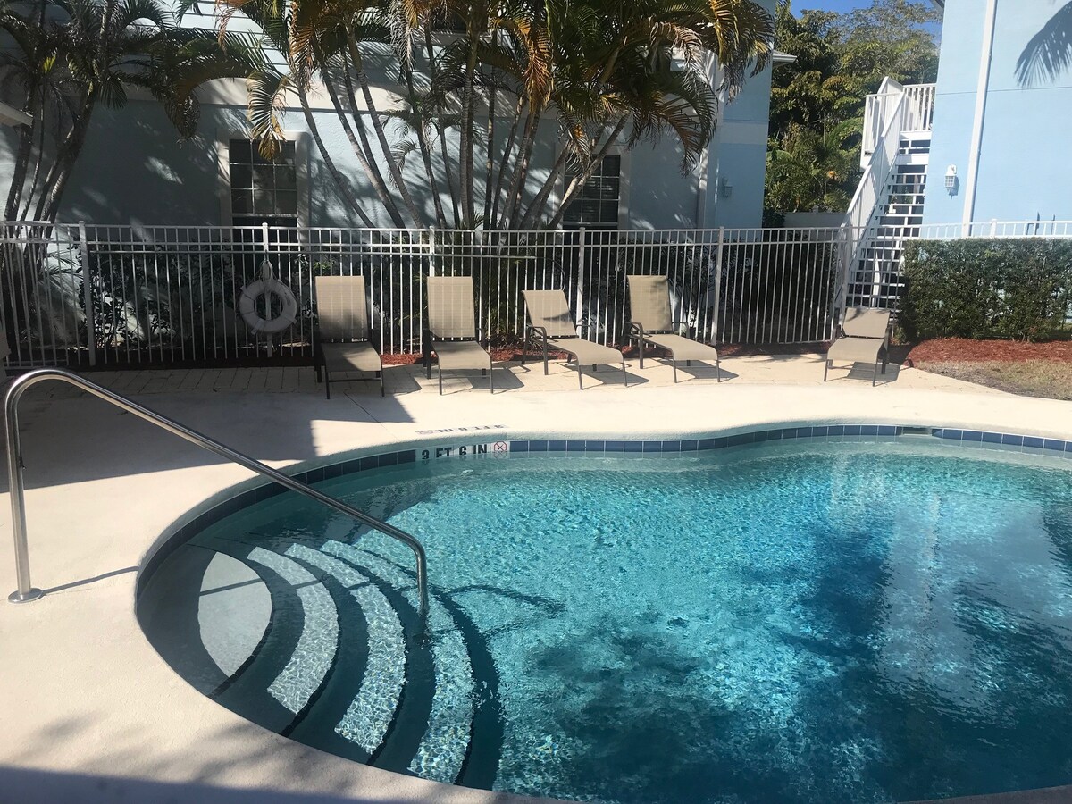 A shared heated pool is presented within a tranquil outdoor area, surrounded by palm trees. Several lounge chairs are positioned along the poolside, offering a relaxing space. The pool features steps leading into the water, creating an inviting atmosphere for enjoying warm days.