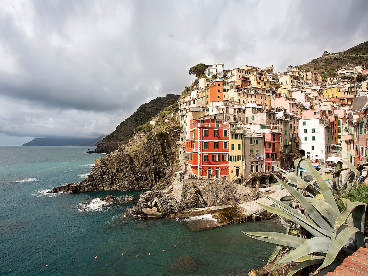 A coastal view of Riomaggiore showcases the charming architecture of colorful buildings ascending the hillside. The rocky shoreline and turquoise waters create a serene environment, while the cliffs and distant coastline add depth to the picturesque landscape under a cloudy sky.