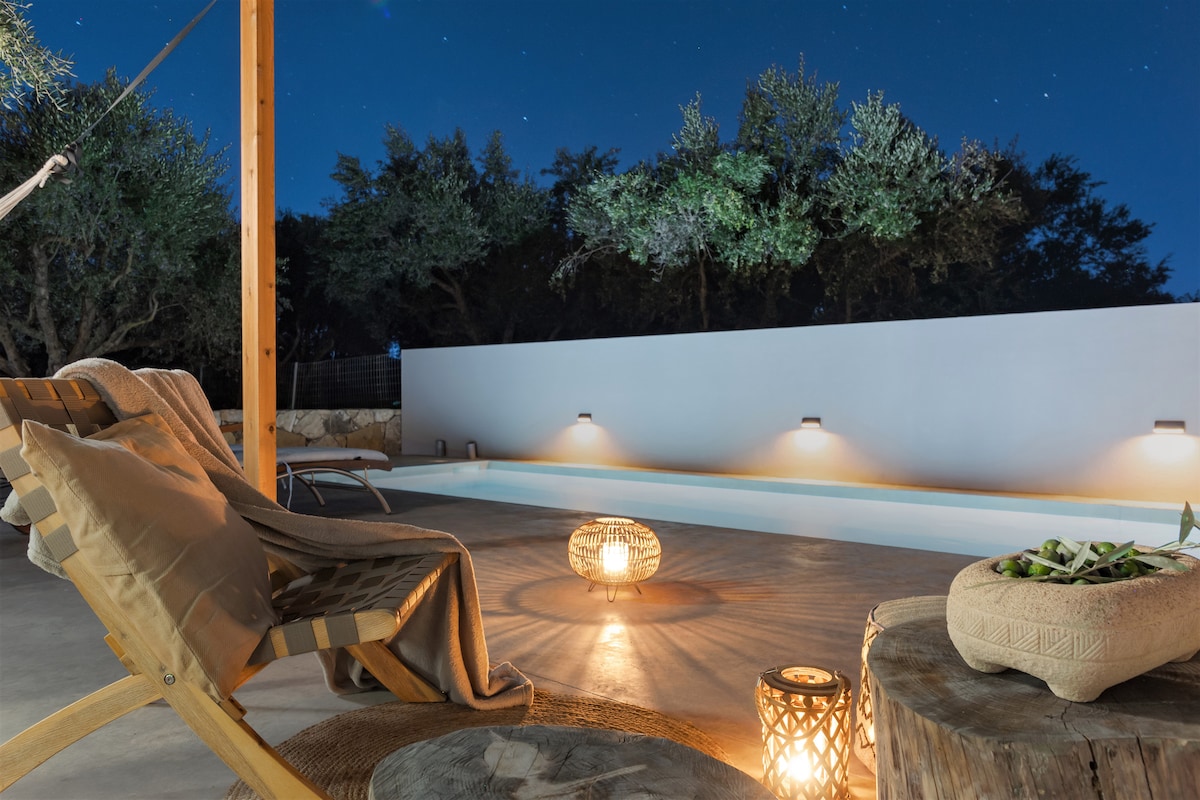 A serene outdoor space features a private pool surrounded by olive trees, enhanced by soft lighting. Two wooden lounge chairs are positioned comfortably nearby, accompanied by woven fixtures that add texture and warmth to the area.