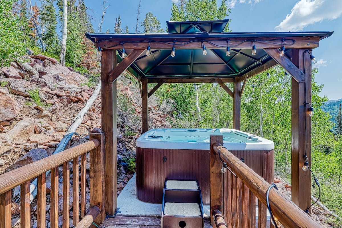 A hot tub is situated under a wooden gazebo in a natural setting, surrounded by trees and rocky terrain. Soft lighting fixtures hang from the structure's ceiling, creating a relaxing ambiance. Access steps lead up to the hot tub area, which offers scenic views of the landscape.