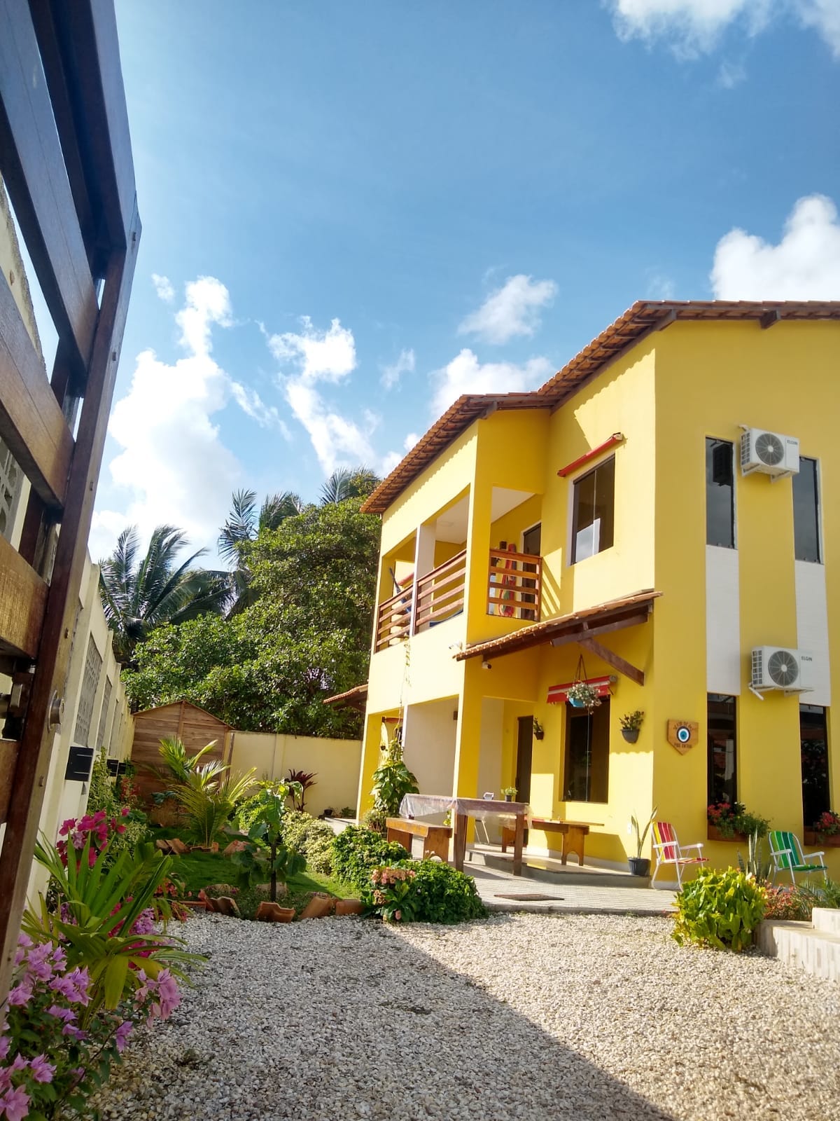 A two-story yellow building is surrounded by lush greenery and colorful flowers. Bright blue skies and scattered clouds form the backdrop. A patio with outdoor seating is visible, enhancing the inviting exterior. Air conditioning units are mounted on the walls, providing comfort.