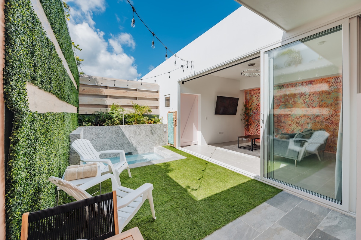 A private oasis terrace captures sunlight, featuring a refreshing plunge pool and a well-maintained grassy area. Lounge chairs are arranged on the terrace, complemented by lush green walls. Sliding glass doors provide a seamless connection to the spacious living area, enhancing the tranquil ambiance.