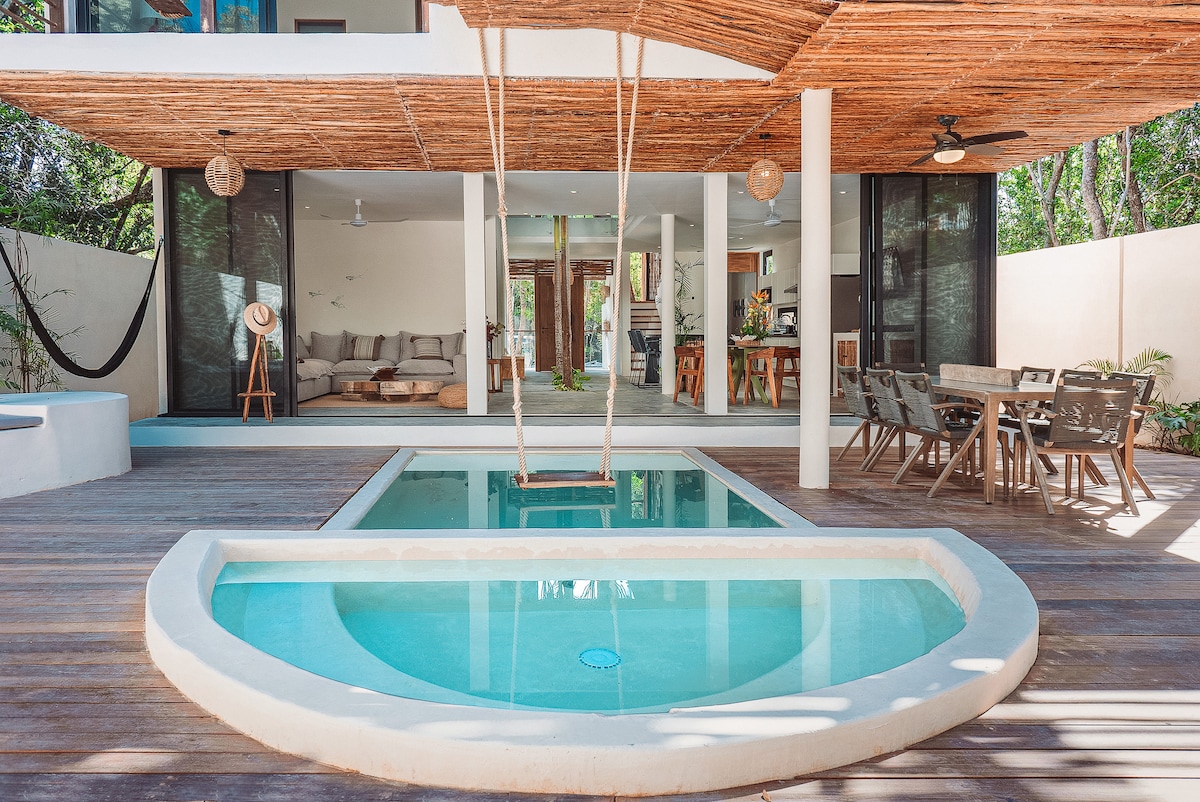 A private outdoor area features a small plunge pool adjacent to a larger pool, both set within wooden decking. A swing hangs from a thatched roof, and views of a spacious indoor area are visible through large glass doors. Outdoor seating is arranged nearby.