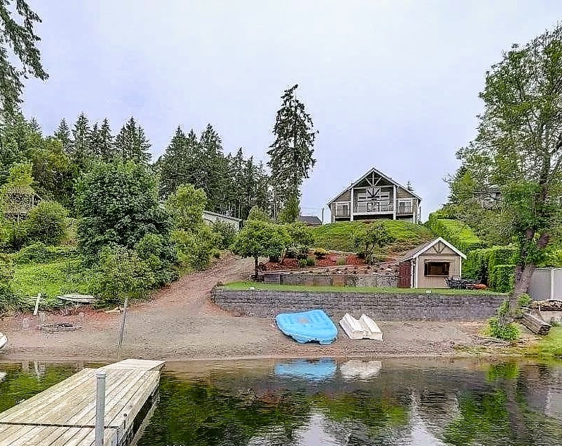 A serene lakeside view is presented, featuring a spacious home set on a gentle slope. The sandy beach blends with lush greenery, while a wooden dock leads into calm waters. Watercraft and beach toys are positioned nearby, inviting outdoor relaxation and activities.