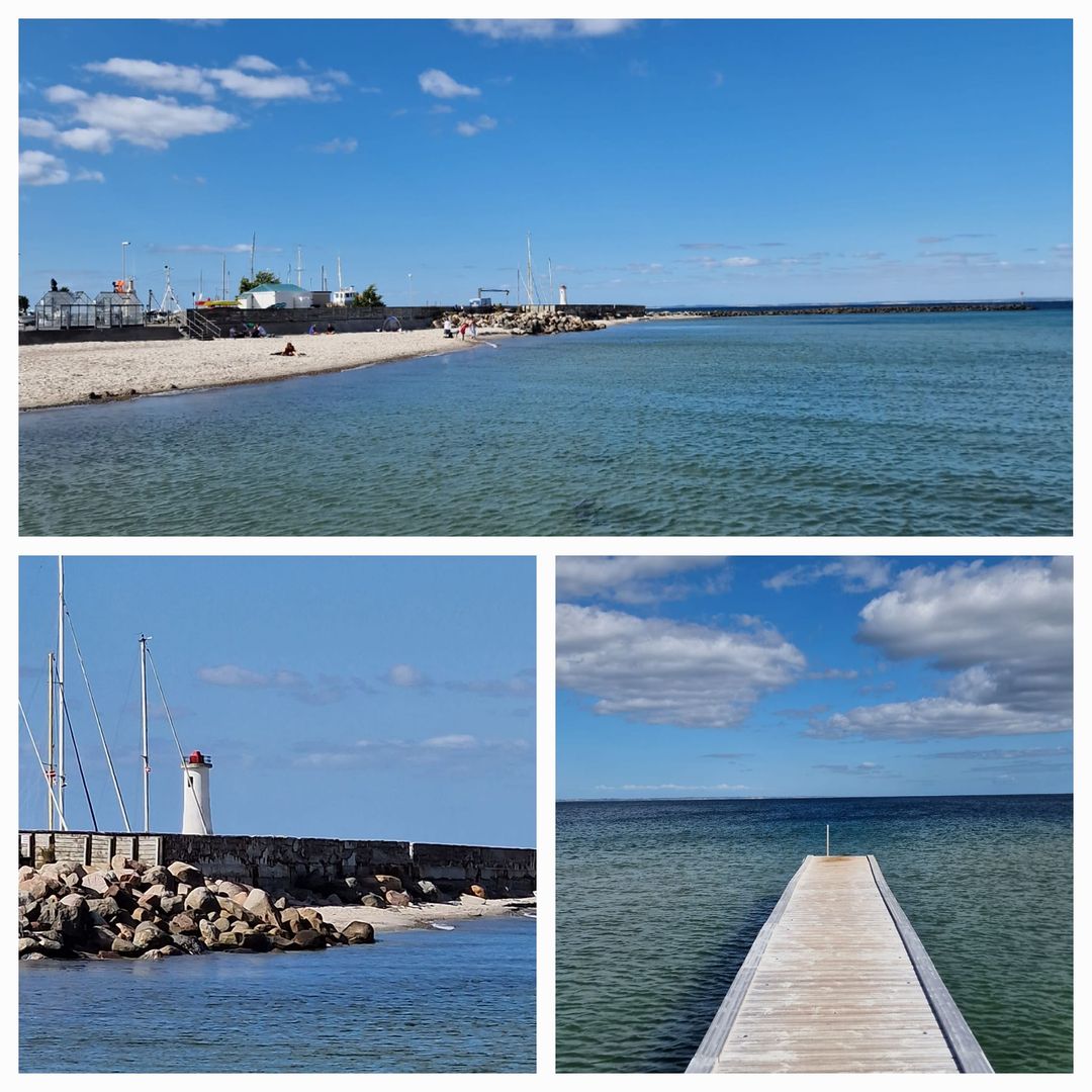 The image showcases a serene coastal view featuring a sandy beach and calm waters. A wooden pier extends into the sea, surrounded by sailboats and a lighthouse. The sky is clear with scattered clouds, adding to the tranquil atmosphere.