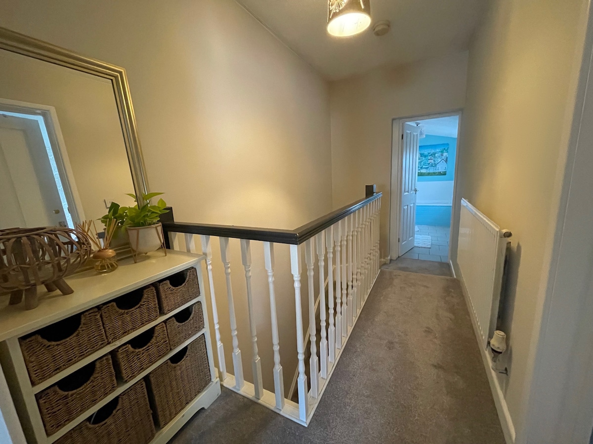 A well-lit hallway features a light-colored wall with a mirror above a storage unit. The unit holds woven baskets and is complemented by a potted plant. A softly carpeted floor leads to an open door at the end, revealing a glimpse of another room.