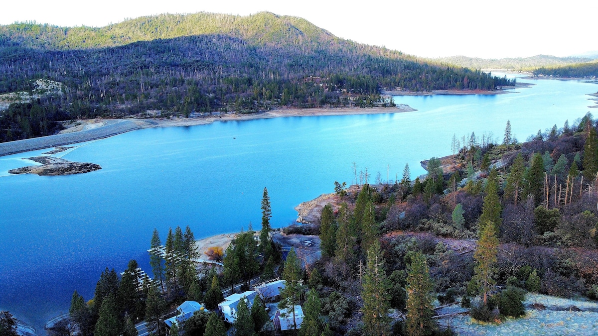 A serene view of a vast lake is framed by rolling hills and trees. The water's surface reflects a clear blue sky, with gentle ripples noticeable. Areas of sandy shoreline and patches of greenery are visible along the lake's edge, creating a tranquil landscape.