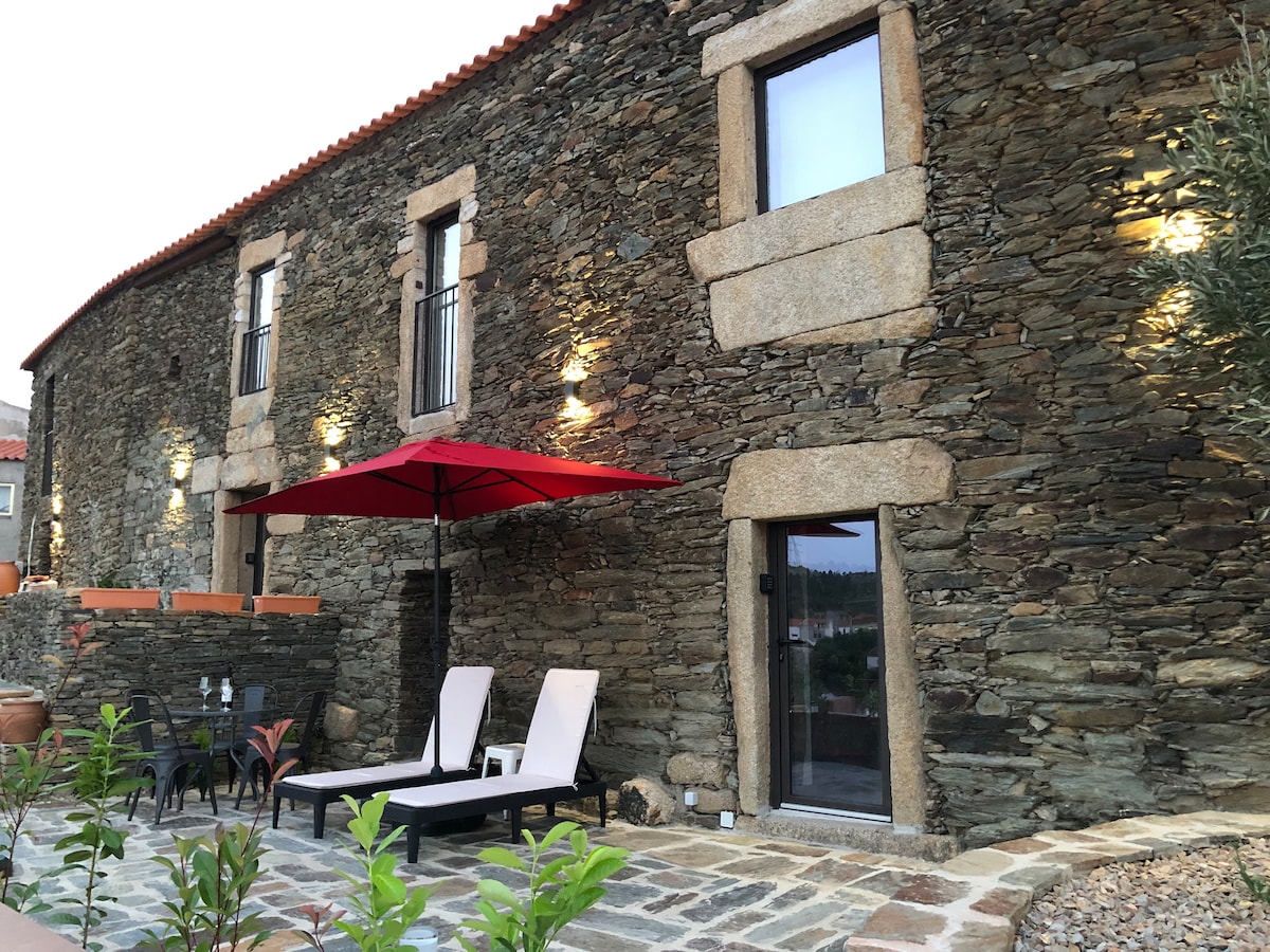 The exterior of the house showcases rustic stone walls under warm evening lighting. A shaded seating area is provided by a red umbrella over two loungers. Paved pathways and greenery enhance the inviting atmosphere of the outdoor space.