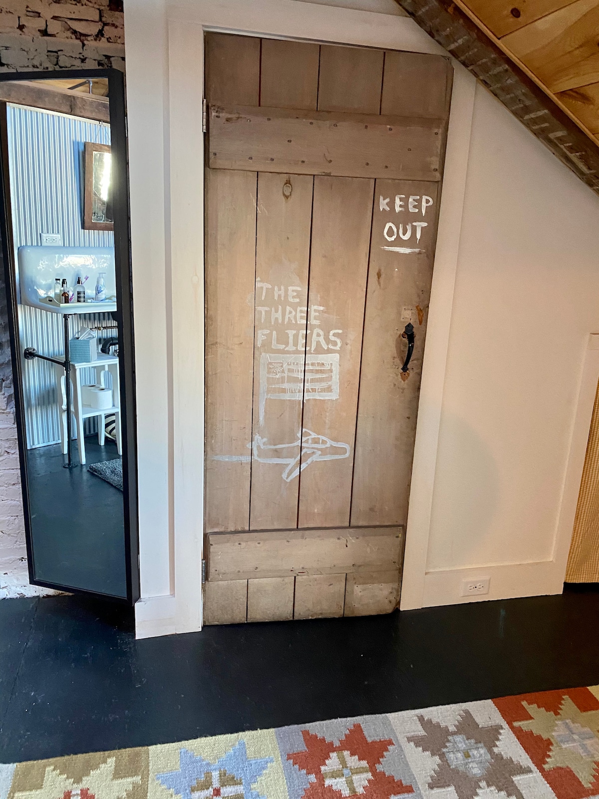 A rustic wooden door features hand-painted text reading 'KEEP OUT' and 'THE THREE FLIRS.' The door is attached to a quaint wall, with a black-framed mirror visible to the left and a glimpse of a warm-toned living area in the background.