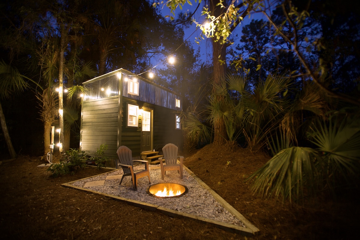 A cozy tiny house is illuminated by string lights, nestled among tall trees. A fire pit sits in front, surrounded by gravel, with two chairs positioned nearby, inviting relaxation in a serene outdoor setting.