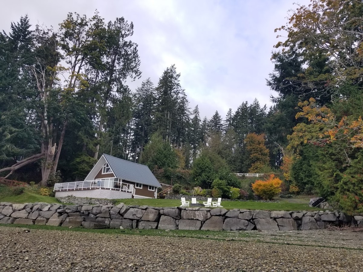 A charming cottage sits among tall trees, framed by a lush landscape. White chairs are arranged on a stone terrace, inviting relaxation. The structure features a gabled roof and a large deck, positioned near the shoreline.