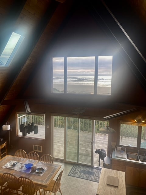 The spacious dining area features a large wooden table set with blue dishware, adjacent to a well-equipped kitchen. Large windows offer an ocean view and allow natural light to fill the room, while a sliding glass door leads to the expansive outdoor deck.