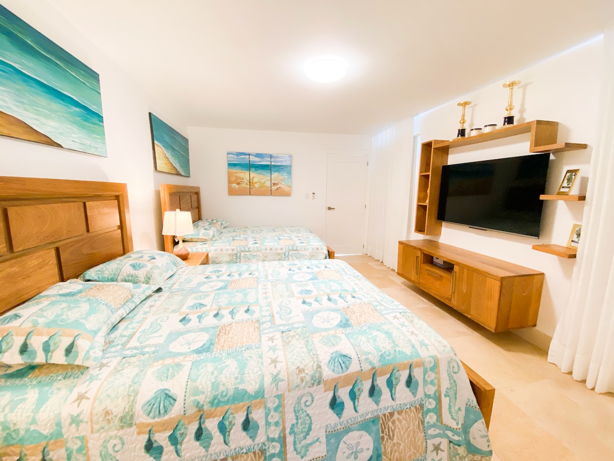 A spacious bedroom features two Queen beds adorned with patterned bedding in soothing shades of blue and green. A large wall-mounted TV is set across from the beds, complemented by a wooden entertainment unit. Artwork depicting beach scenes enhances the room's coastal theme.