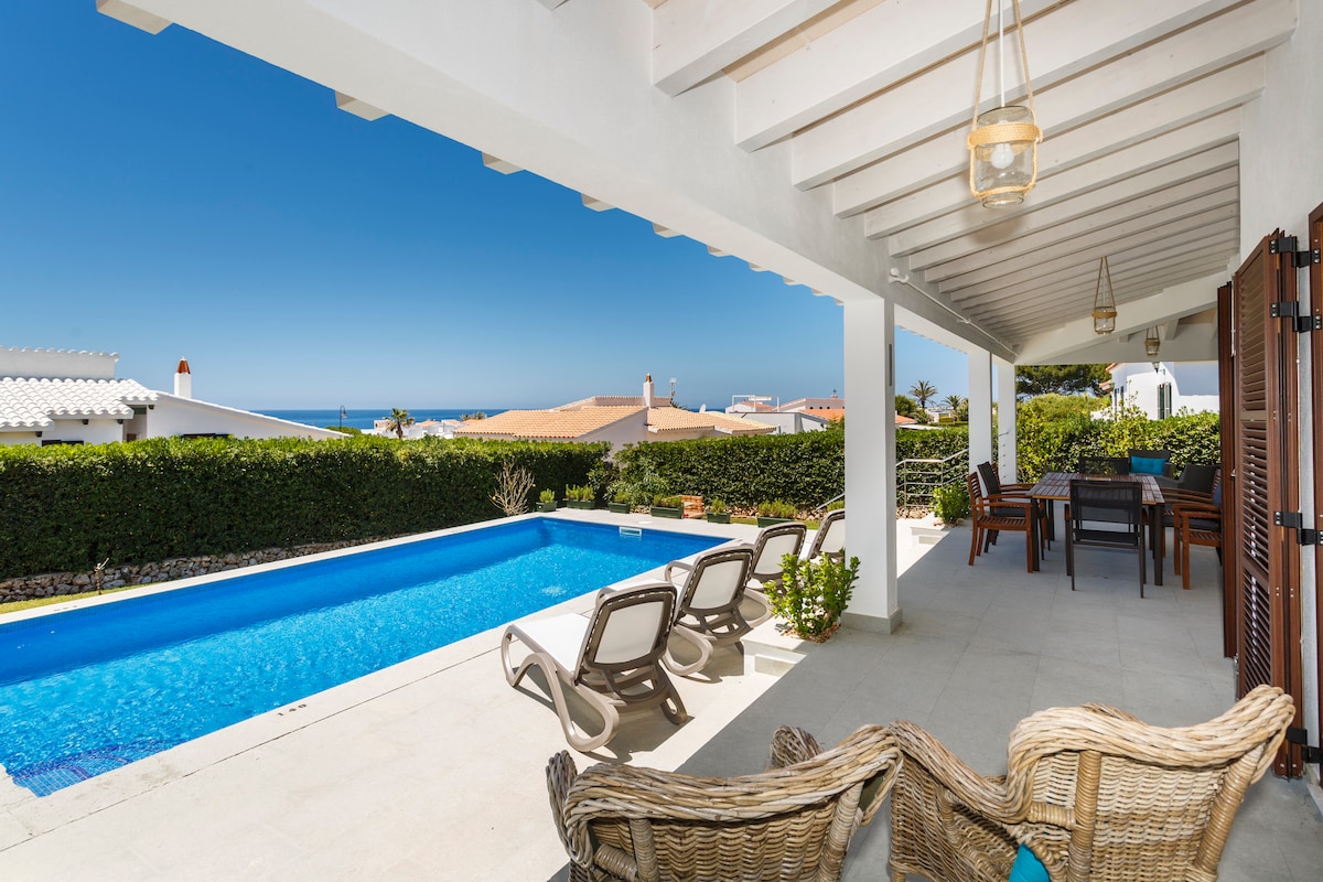 A spacious terrace features comfortable seating and a dining area, overlooking a private swimming pool. The inviting blue water of the pool reflects the clear sky, while greenery frames the scene, enhancing the outdoor relaxation space.