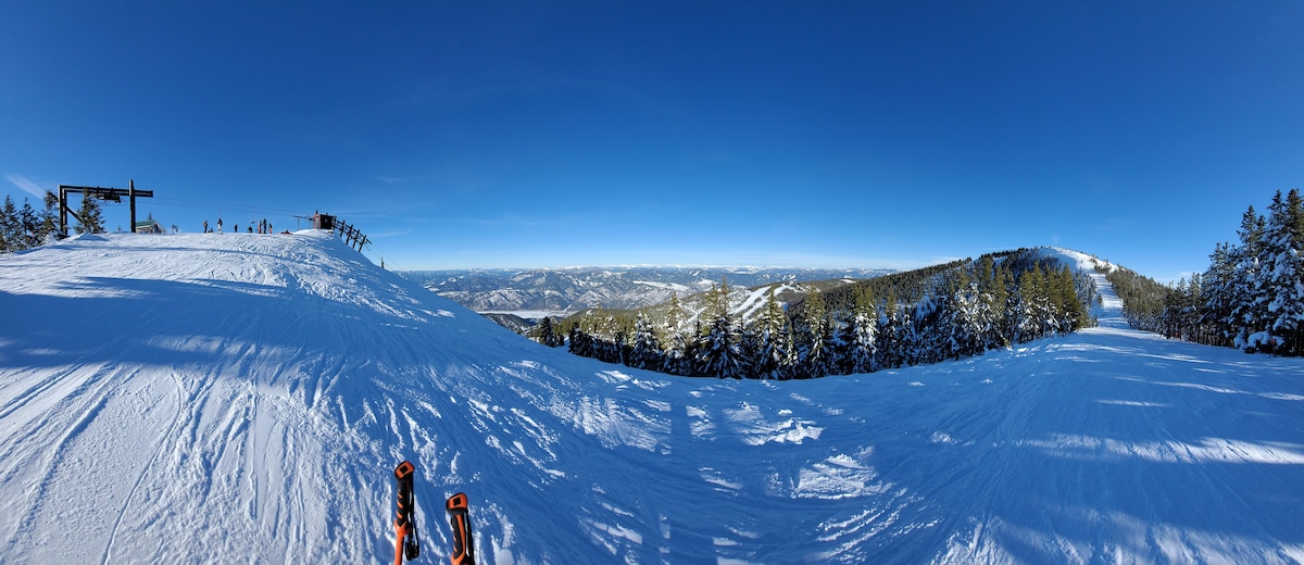 A wide, snow-covered slope is visible under a bright blue sky. The landscape features softly rolling hills and distant mountains, with a group of people enjoying various winter activities at the horizon. Multiple ski tracks are imprinted in the snow, leading down the gentle incline.